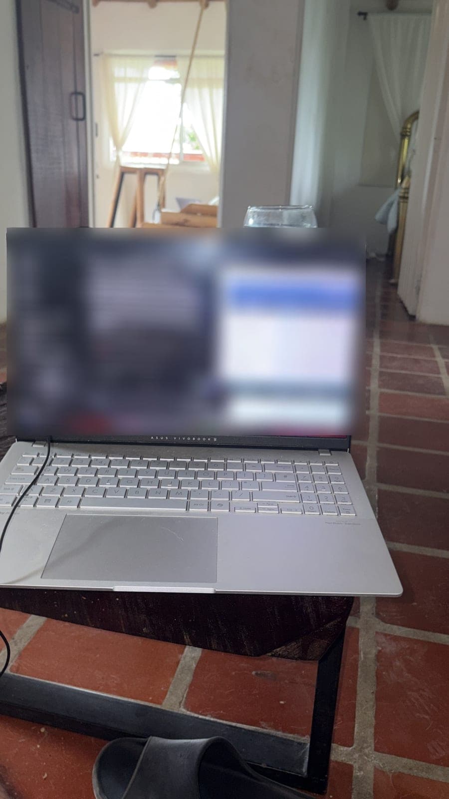 A laptop on a small table in a room with a brick floor, its screen blurred